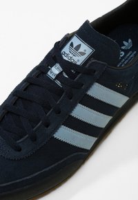 Mörkblå snörskor i mocka med tre ljusblå ränder, svart yttersula och en grå tungetikett med Adidas-logotypen. Skosnörena är mörkblå.