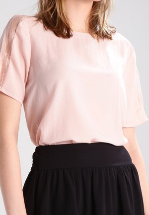 Vrouw draagt een lichtroze blouse met kanten mouwen, gestopt in een zwarte rok met hoge taille, zichtbaar van schouders tot taille.