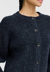 Tmavě námořnický žebrovaný pletený cardigan s kulatým výstřihem, který má pět velkých černých knoflíků a texturované detaily na rukávech.