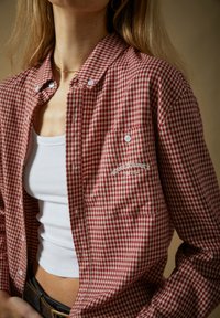 Γυναίκα που φοράει κόκκινο και λευκό καρό πουκάμισο με κουμπιά πάνω από λευκό λεπτό ριπτό τοπ, με τσέπη στο στήθος που φέρει την επιγραφή "House of Sunny".