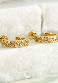 Guldörhängen i form av ringar med en texturerad, vävd design och satta med små klara stenar. Arrangerade på en ljus marmor yta.