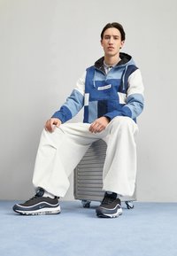 Sudadera de mezclilla patchwork en azul y blanco con cremallera, combinada con pantalones de pierna ancha blancos y zapatillas Nike Air Max 97 negras.