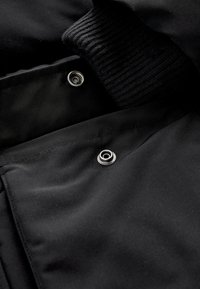 Gros plan sur un tissu de veste noire avec des boutons pression argentés et un poignet en tricot côtelé à l’intérieur de la manche.