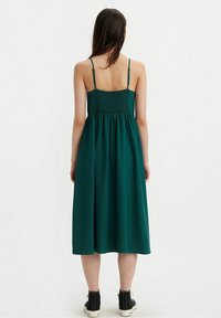 Grünes ärmelloses Kleid mit gerafftem Rücken, fließendem Rock und knöchellanger Schnitt. Getragen mit schwarzen High-Top-Sneakern.