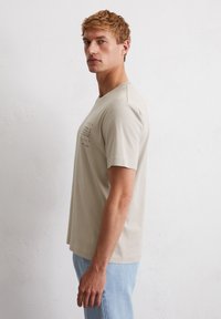 Light beige cotton t-shirt with short sleeves and a crew neckline. Front features printed text in black. Paired with light blue denim jeans.