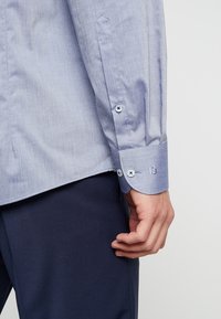 Close-up van de linkerhand van een persoon die naast hun lichaam rust, gekleed in een lichtblauwe, lange mouwen, knoopshirt en donkerblauwe broek.