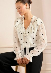 White blouse with black heart patterns, ruffled collar and cuffs, V-neck design, and thin tie detail; paired with black high-waisted pants.