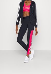 La ropa deportiva Adidas incluye una sudadera con capucha azul marino y cremallera, un sujetador deportivo rosa y leggings de talle alto con un detalle rosa y el logo. Zapatillas blancas completan el conjunto.