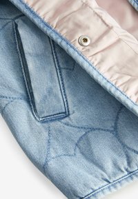 Veste en denim avec un extérieur matelassé bleu clair à motifs ronds et une doublure en satin rose, dotée d'une fermeture à pression et d'une poche intérieure.