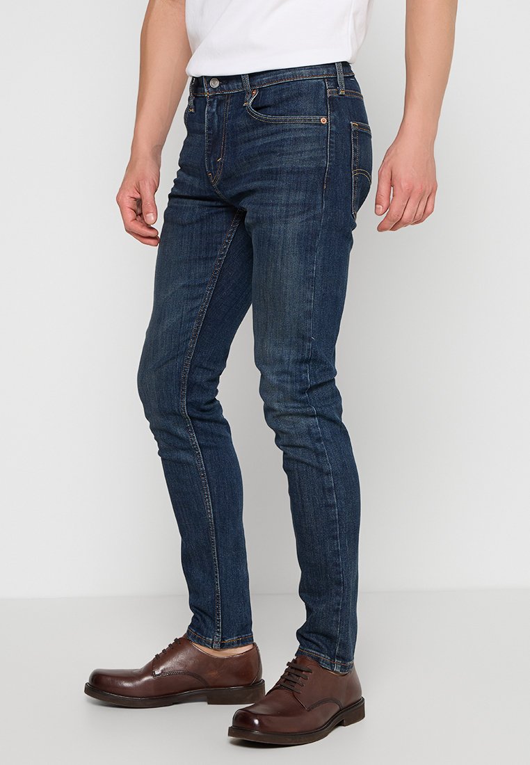 Dżinsy o ciemnym odcieniu, slim fit, z pięcioma kieszeniami i pomarańczowymi szwami, zestawione z brązowymi skórzanymi butami.