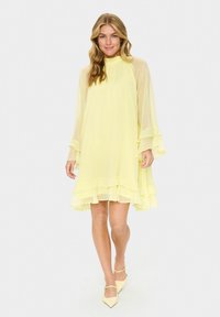 Short yellow dress with a high neckline, flowing fabric, layered ruffles at sleeves and hem, and sheer detailing. Paired with matching light shoes.