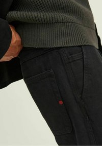 Pantalones negros con un bolsillo lateral que presenta un detalle de punto rojo, combinados con un suéter acanalado verde oscuro, enfatizando un look casual en capas.