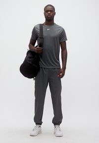 Άνδρας που φοράει σκούρο γκρι αθλητικό σύνολο και λευκά αθλητικά παπούτσια, κρατώντας μια μαύρη τσάντα duffel στον ώμο, στέκεται μπροστά σε μια απλή φόντο.