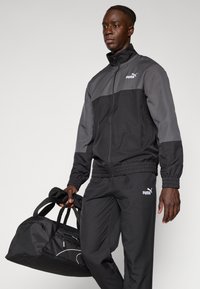 Schwarzer und grauer Sport-Trainingsanzug mit einer Reißverschlussjacke und elastischen Bündchen, kombiniert mit einer schwarzen Sporttasche mit weißen Akzenten.
