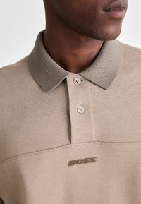 Beige polo shirt with a ribbed collar, featuring two buttons and a subtle embossed logo. Soft texture and clean design.