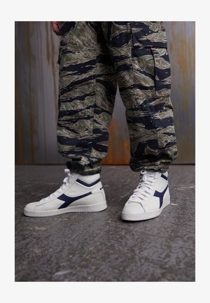 Sneakers alte bianche con accenti blu navy, pelle texturizzata e suola in gomma. Pantaloni cargo in verde e nero a motivo mimetico.