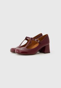 Scarpe col tacco in pelle verniciata bordeaux con T-strap, dettagli perforati sulla punta e tacco spesso. Texture liscia e design classico.