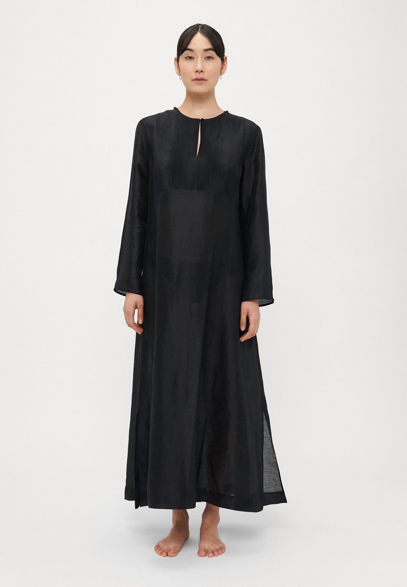 Rochie neagră cu mâneci lungi, cu partea superioară plisată, decolteu rotund, detaliu frontal cu deschidere în formă de cheie și fante laterale, realizată din țesătură ușoară și transparentă.