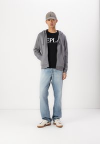 Sudadera gris con capucha y bolsillos frontales, camiseta negra con texto blanco, jeans celestes, y zapatillas beige con detalles oscuros.