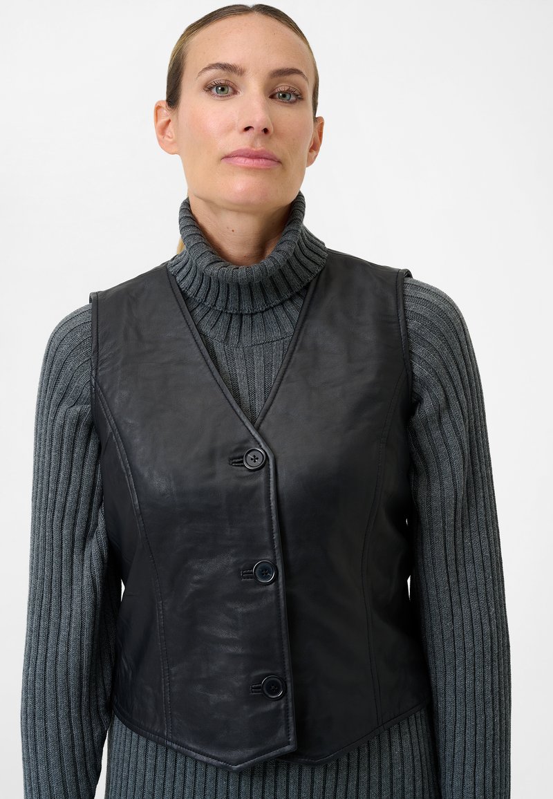 Schwarze Lederweste mit figurbetontem Design, V-Ausschnitt, drei Knöpfen und glatter Textur, getragen über einem grauen, gerippten Rollkragenpullover.