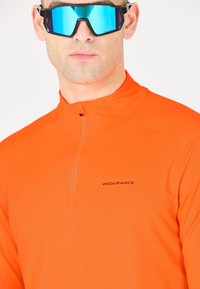 Klar orange långärmad topp med halvdragkedja och "ENDURANCE"-logotyp. Har ett glatt, elastiskt material. Bärs med blå spegelglasögon.