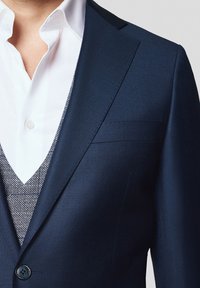 Granatowa marynarka o fakturowanym wykończeniu, z wyłogami, zapinana na jeden guzik, założona na biały koszulę i wzorzystą kamizelkę.