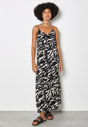 Frau mit lockigem Haar, die ein schwarz-weiß gemustertes Maxikleid und schwarze Sandalen trägt, steht vor einer schlichten hellen Wand.