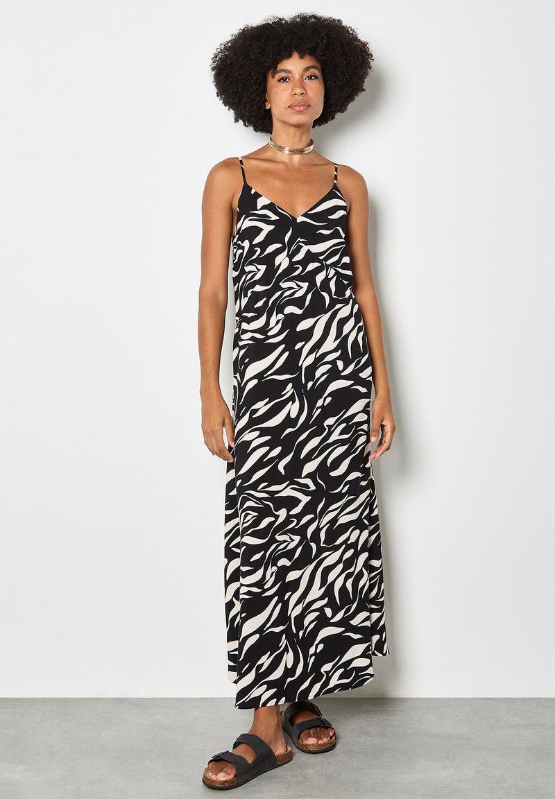Kobieta o kręconych włosach, ubrana w czarno-białą, wzorzystą sukienkę maxi i czarne sandały, stoi na tle gładkiej jasnej ściany.