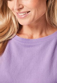 Lila, weiches Stoffoberteil mit weitem Halsausschnitt, das eine glatte Textur zeigt. Das Bild zeigt teilweise sichtbares, blondes Haar.