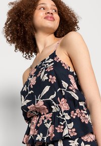 Top peplum floral com fundo azul-marinho, apresentando padrões de flores em rosa claro e creme. Alças finas e textura de tecido suave e fluida.