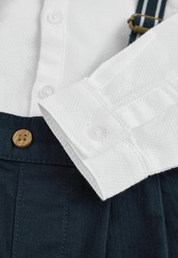 Beli teksturirani ovratnik s srajco z gumbi poleg temno modrega platna z gubami in lesenim gumbom, ki ima naramnice s črtastim vzorcem.