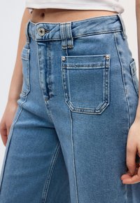 Γυναίκα που φοράει ψηλόμεσα μπλε τζιν με μεγάλες μπροστινές τσέπες και λευκό κολλητό μπλουζάκι, με το χέρι ακουμπισμένο κοντά στον μηρό.