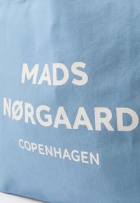 Jasnoniebieska torba materiałowa z napisem "MADS NØRGAARD COPENHAGEN" czcionką pogrubioną w białym kolorze. Gładka faktura, prosty design i brak dodatkowych wzorów.