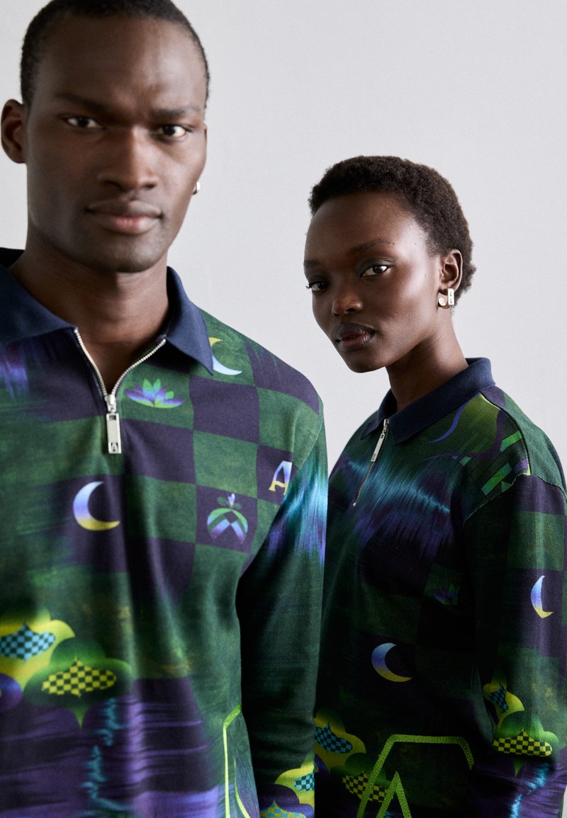 Grønne zip-up skjorter med blå og lilla mønstre, der fremviser geometriske figurer og symboler. Blødt stof med en struktureret overflade.