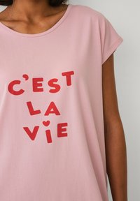 Lys rosa T-shirt med korte ærmer og rød trykt tekst "C'est la vie" i forskellige fede skrifttyper, med en afslappet løs pasform og blød tekstur.