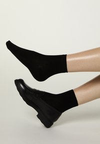 Schwarze gerippte Socken, getragen mit schwarzen Leder-Loafers, mit runder Zehenpartie und dicker Gummisohle. Beine teilweise sichtbar vor neutralem Hintergrund.