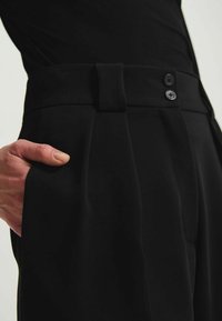 Pantalons noirs taille haute avec plis, dotés d'une large ceinture et d'une double fermeture boutonnée. Tissu lisse avec poches latérales.