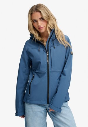 Lyshåret kvinde iført blå hættejakke med sort lynlås og lyseblå jeans, stående mod en hvid baggrund.