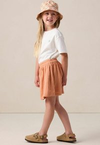 Weißes Baumwoll-T-Shirt mit grüner Stickerei, orangefarbene strukturierte Rock mit rosa floralen Akzenten und braune Sandalen mit Schnallen. Bucket Hat im Muster.