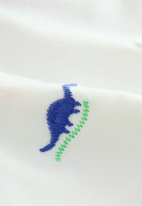 Tissu blanc avec un motif de dinosaure brodé en bleu et des accents en zigzag vert, présentant une texture douce et des détails clairs.