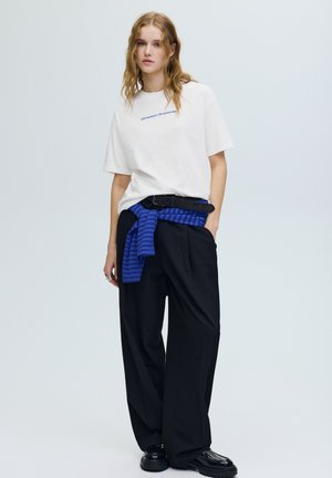 Wit katoenen T-shirt met blauwe tekst, zwarte wijde broek met rechte pijpen en een blauw gestreepte trui gestrikt om de taille. Zwarte veterlaarzen.