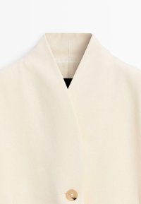 Beige frakke med en bred krave og en enkelt tan-farvet knap, vist tæt på den øverste forside mod en hvid baggrund.