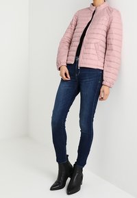 Chaqueta bomber acolchada de color rosa claro con cierre de cremallera, combinada con unos vaqueros ajustados azul oscuro y botas negras de tobillo. Textura suave y diseño ajustado.