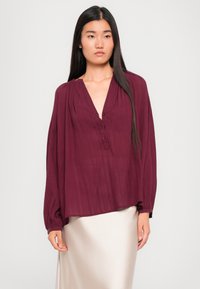 Femme à cheveux longs et foncés portant un blouse texturée maroon ample et une jupe satin beige clair, se tenant devant un fond uni.
