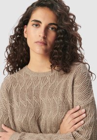 Beige gestrickter Pullover mit Wellenmuster und offener Stricktechnik, Rundhalsausschnitt und langen Ärmeln. Die Textur wirkt leicht und atmungsaktiv.