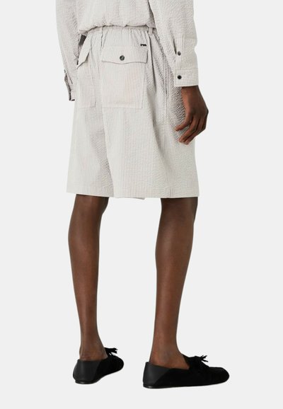 Pantaloni corti a righe, di colore chiaro e dalla vestibilità ampia, dotati di tasche laterali e una tasca posteriore con bottone. Indossati con scarpe nere.