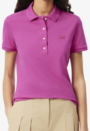 Femme portant un polo à manches courtes rose vif avec des boutons blancs et un pantalon beige plissé, les mains dans les poches.