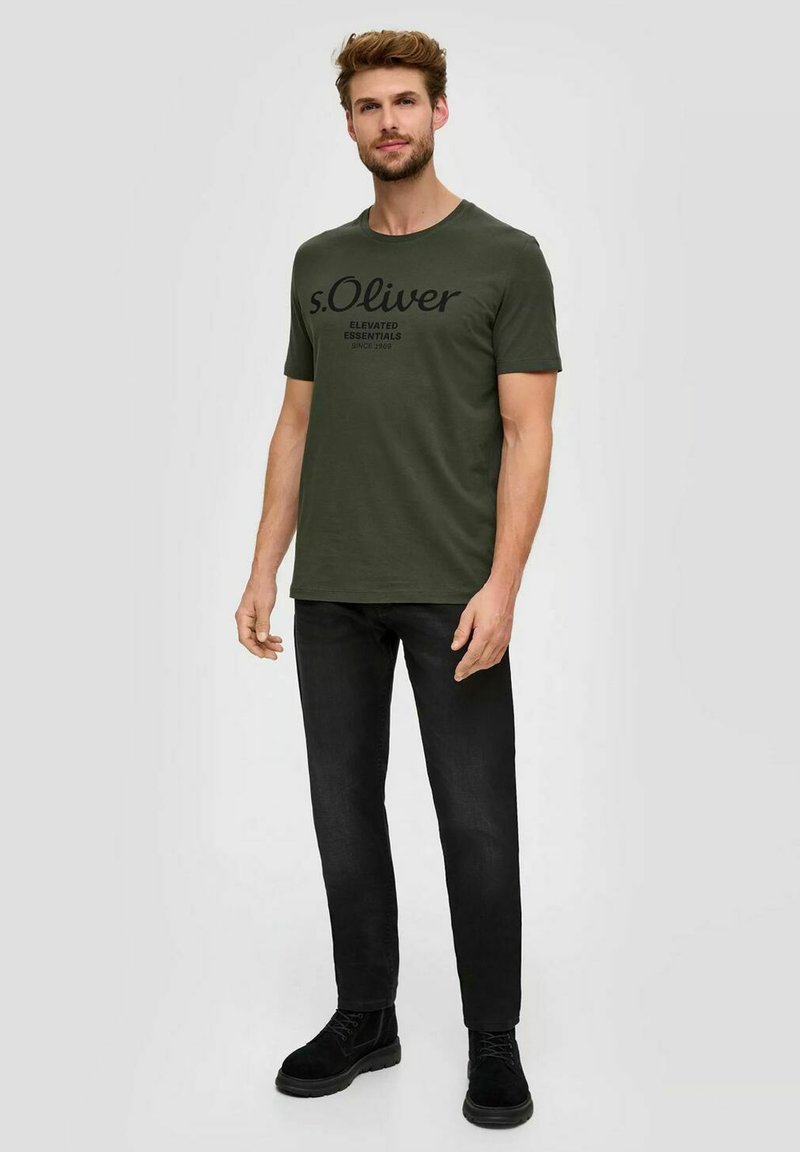 Olivgrünes T-Shirt mit schwarzem Text "s.Oliver ELEVATED ESSENTIALS" auf der Vorderseite, kombiniert mit schwarzen Slim-Fit-Jeans und schwarzen Stiefeln.