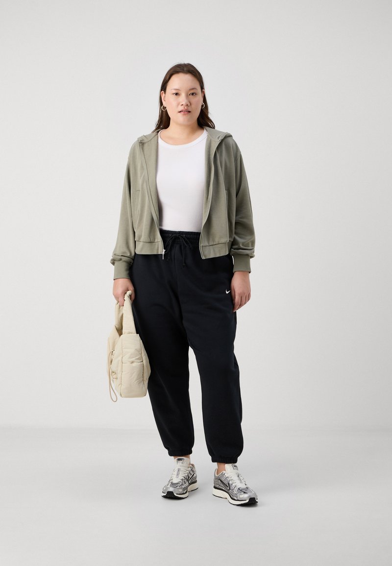 Sudadera corta de color verde oliva, camiseta ajustada blanca, pantalones de chándal negros con puños elásticos, zapatillas plateadas y un bolso acolchado beige.