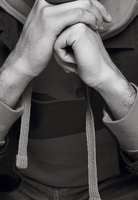 Zbliżenie na splecione dłonie. Dłonie są wyraźne, z jasnym owłosieniem, a tekstura jasnoszarej tkaniny z ozdobnym sznurkiem jest widoczna.